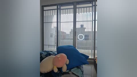 Media: Video of a cozy bedroom with a plush white and pink toy bear on a bed with blue and green sheets, facing a large window with vertical blinds and a circular moon-shaped light.