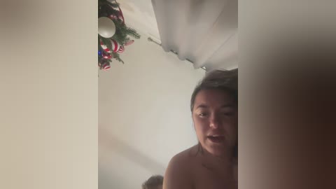 Media: Video of a young, bare-chested woman with dark hair, looking concerned, in a dimly lit room with a misty ceiling. A festive wreath with red and green elements hangs on the wall.