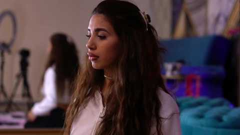 Media: Video of a young woman with long, wavy brown hair, wearing a white blouse, standing in a dimly lit room with blurred figures in the background.