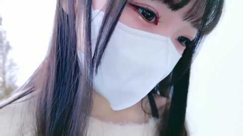 Media: Close-up video of an East Asian woman with long, straight black hair, wearing a white face mask, red eye makeup, and a beige top. Background is blurred, showing a cloudy sky.