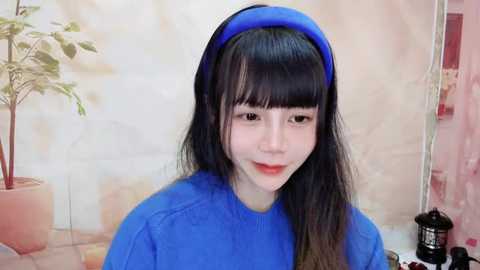 Media: Video of a young Asian woman with long black hair and bangs, wearing a bright blue headband and matching blue top. Background features a pale beige wall, potted plant, and a black kettle.