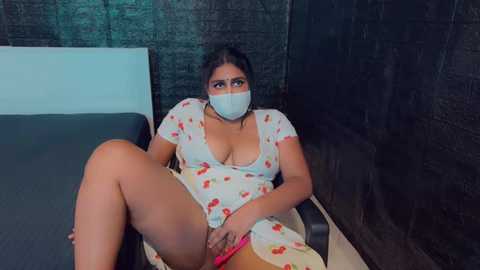 Media: Video of a South Asian woman with medium skin tone, dark hair, wearing a white dress with red cherries, a mask, and a pink vibrator, sitting on a black couch in a dimly lit room with dark brick walls.