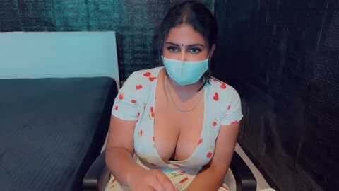 Media: Video of a South Asian woman with dark hair, fair skin, wearing a floral dress, a blue surgical mask, and sitting in a wheelchair. She has a large bust and is in a dimly lit room with dark walls and a bed.