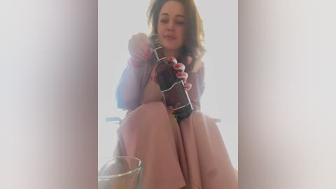 Media: A video of a young woman with fair skin, dark hair, and a light pink dress, sitting in a bright, minimalist room, holding a glass bottle of soda.