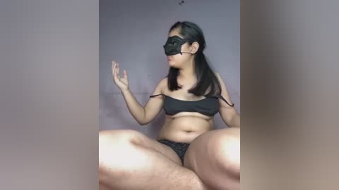 Media: Video of a plus-sized Asian woman with long black hair, wearing a black bra and panties, sitting cross-legged on a floor, with a black blindfold and mask covering her eyes.