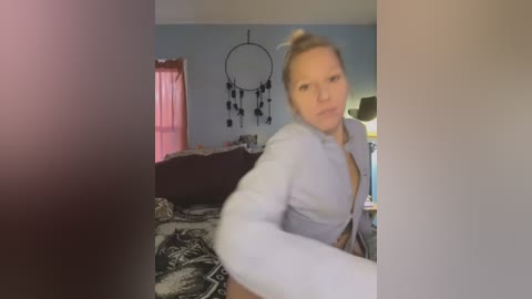 Media: A video of a blonde woman in a light blue shirt, leaning slightly forward, taken in a bedroom with a dreamcatcher on the wall, a bed with a black-and-white patterned blanket, and a pink curtain.