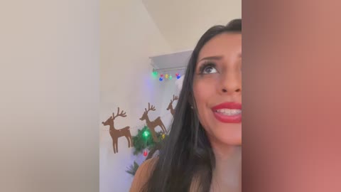 Media: Video of a smiling, light-skinned woman with long black hair, wearing red lipstick, standing next to a white wall adorned with reindeer cutouts and Christmas lights.