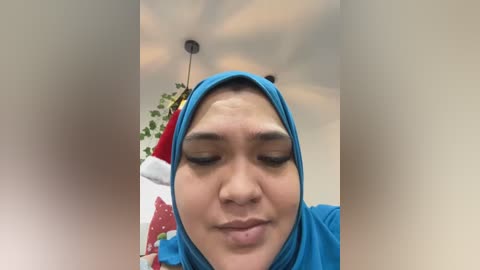 A close-up video of a woman with light brown skin, wearing a blue hijab, smiling, with a blurred background featuring a Christmas tree and greenery.