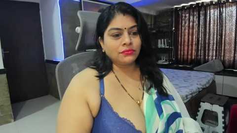 Media: Video of a South Asian woman with dark hair, fair skin, and a slender build, wearing a blue bra, seated in a modern bedroom with a grey upholstered chair, dark curtains, and a neatly made bed.