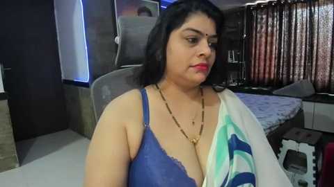 Media: A video of a plus-size South Asian woman with medium skin tone, black hair, and a blue bra, sitting in a modern bedroom with a white bed, brown curtains, and gray chair.