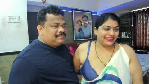 Media: Video of an overweight South Asian couple, smiling, in a modern living room with framed family photos, dark wooden furniture, and a bed.