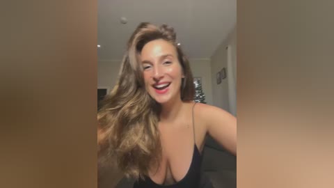 Media: Video of a smiling, light-skinned woman with long, wavy brown hair, wearing a black spaghetti-strap dress, in a modern living room with beige walls and recessed lighting.