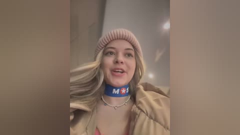 Video of a smiling young woman with long, blonde hair, wearing a pink beanie, blue \"M\" necklace, and a beige jacket.