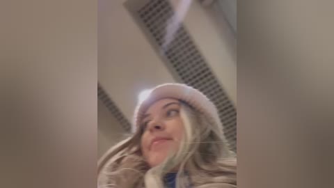 A video of a woman with long blonde hair and a beige hat, looking upward in a room with a vent and ceiling lights.