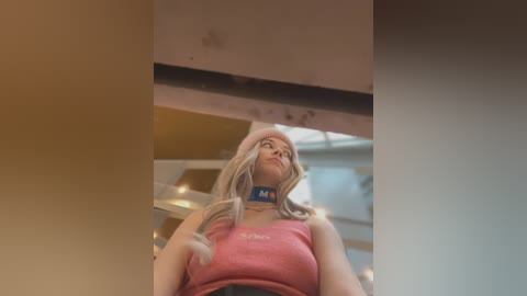 Media: Video of a young blonde woman with a pink beanie, wearing a pink tank top, standing inside a dimly lit, modern indoor space.