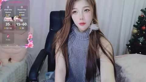 Media: Video of a young Asian woman with long brown hair, wearing a gray turtleneck sweater, sitting on a black chair in a cozy room with a Christmas tree and digital game interface overlay.