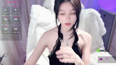 Media: A video of an East Asian woman with long black hair, wearing black lingerie with lace trim, sitting on a white bed with fluffy pillows.