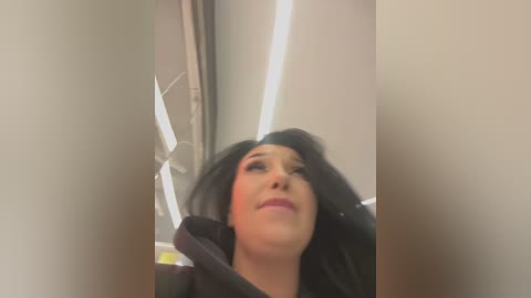 Media: A video of a woman with long black hair, wearing a dark jacket, looking up, possibly in a public restroom, with fluorescent lighting and beige walls in the background.