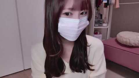 Media: Video of a young Asian woman with long, straight black hair and light skin, wearing a white surgical mask, sitting in a room with a pink bed and white wardrobe.