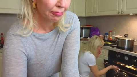 Media: Video of a blonde woman in a light grey ribbed sweater, smiling, and another blonde woman in a white t-shirt, cooking in a modern kitchen with beige cabinets and stainless steel appliances.