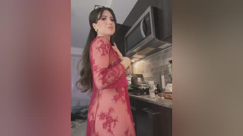 Media: Video of a woman with long dark hair, wearing a sheer red lace dress, standing in a modern kitchen with dark cabinets and a microwave.