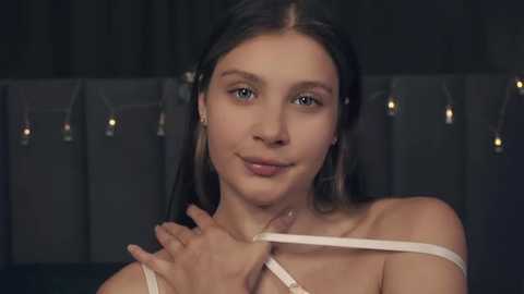 Media: Video of a young woman with light skin and brown hair, wearing a white bra, adjusting the straps. She has a soft smile. Background features a dark curtain with hanging string lights.