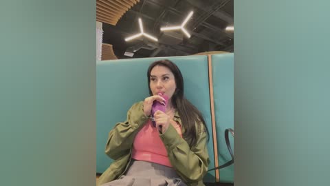 Media: A video of a young woman with long black hair, wearing a green jacket over a pink top and gray skirt, drinking from a pink cup, seated in a teal booth with wooden accents and exposed ceiling beams in a modern cafe.