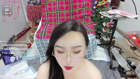 Media: A video shows an Asian woman with long black hair, wearing glasses, seated in a room with a festive, decorated Christmas tree, a red plaid pillow, and a white couch.