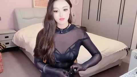 Media: Video of an East Asian woman with long, wavy black hair, wearing a sheer black bodysuit, standing in a modern, minimalist bedroom with a white bed and grey wardrobe.