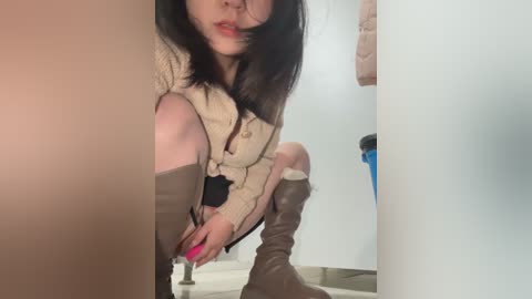 Media: Video of an Asian woman with long black hair, fair skin, and red lipstick, wearing a beige knit sweater, black bra, and thigh-high brown boots, squatting in a bathroom with white walls and a blue trash bin.