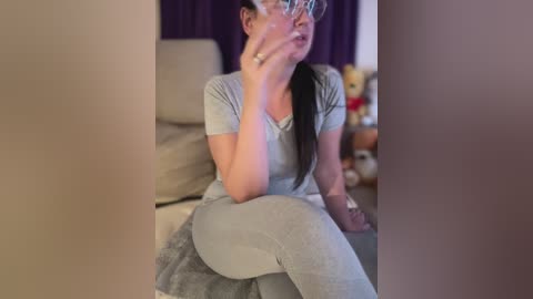 Media: Video of a young woman with glasses, seated on a sofa, wearing grey pajamas, looking thoughtful. Background features a plush teddy bear and purple curtains.