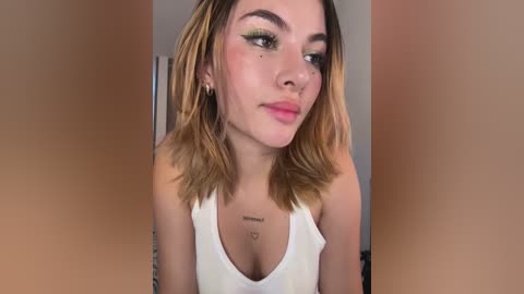 Video of a fair-skinned woman with shoulder-length, ombre hair, wearing a white tank top, and makeup with winged eyeliner. Background is a blurred, peach-colored wall.