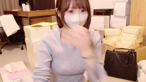 Media: Video of an Asian woman in a gray sweater, wearing a face mask, sitting amidst stacks of yellow file boxes and papers in a cluttered office.