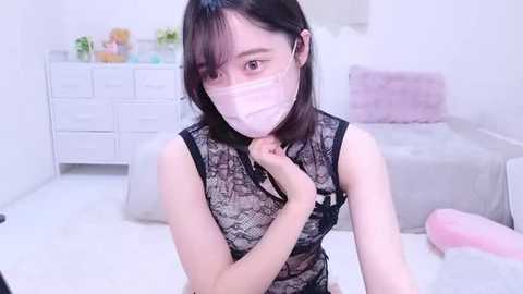 Media: Video of an Asian woman with light skin, wearing a black lace top, pink face mask, and headband. She is seated on a white bed with a pink blanket in a bright, minimalist bedroom.