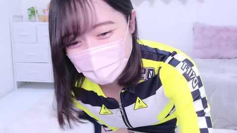 Media: Video of an Asian woman in a bright yellow and black checkered jacket, wearing a pink surgical mask, leaning forward with a slight smile, in a white room with a dresser and bed.