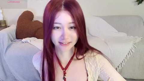 Media: Video of a young Asian woman with long, straight, red hair, fair skin, and small breasts, smiling. She's wearing a white lace top and a red necklace. She's sitting on a beige couch with a brown pillow in a softly lit room.