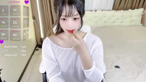 Media: Video of an Asian woman with light skin and long black hair, wearing a white off-shoulder top, licking a red lollipop in a modern bedroom.