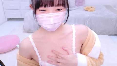 Media: Video of an Asian woman with light skin and black hair, wearing a pink mask and a white lace bra. She is topless, with one hand covering her breast. Background shows a bed with grey sheets and a white dresser.