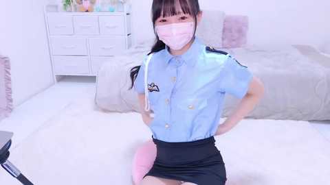 Media: Video of an Asian woman in a blue police uniform, mask, and skirt, kneeling on a white carpet in a minimalist bedroom with light furniture and potted plants.