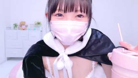 Media: A video of an Asian woman with dark hair in pigtails, wearing a white face mask, black velvet cape, and white lace lingerie, holding a pink drink, in a white room with a dresser.