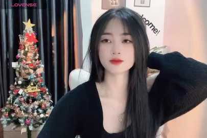 Media: Video of a young East Asian woman with long black hair, fair skin, and red lipstick, wearing a black sweater, standing in front of a decorated Christmas tree in a cozy room.