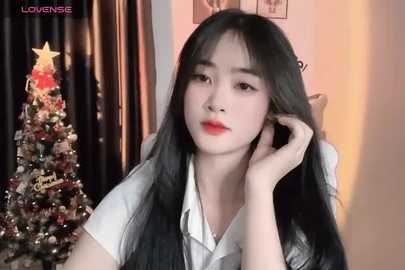 Media: Video of a young East Asian woman with long black hair, fair skin, and red lipstick, wearing a white shirt, posing in front of a decorated Christmas tree with a star.