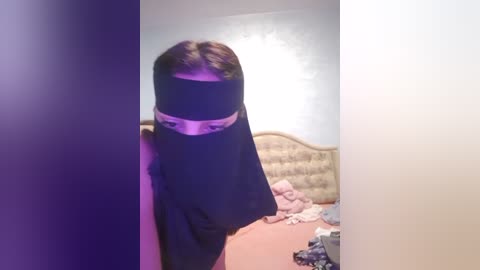 Media: Video of a person with short brown hair wearing a black niqab in a messy bedroom with a beige headboard, scattered clothes, and a pink blanket.