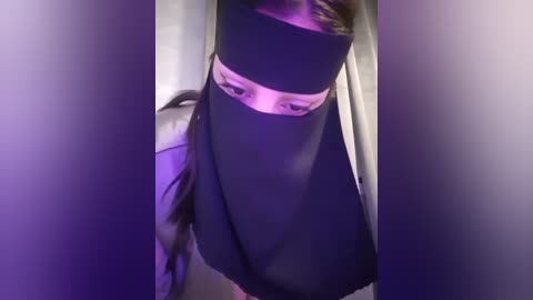 Media: Video of a person wearing a black burqa with a white veil covering the eyes, standing in a dimly lit room with purple lighting. The person's face is not visible.