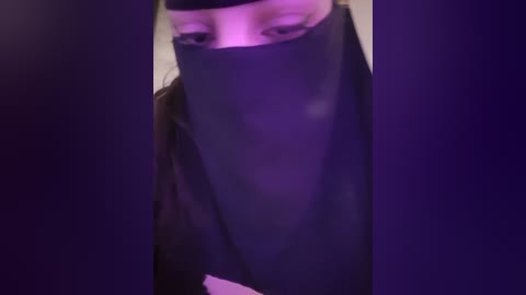 Media: A close-up video of a person wearing a black mask with a slit for the eyes, set against a dark purple background. The person's eyes and nose are visible through the slit.