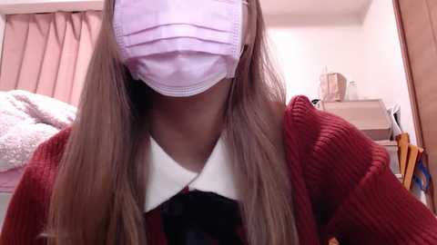 Media: Video of a woman wearing a pink face mask, red cardigan, and white collared shirt, sitting in a bedroom with a bed, wooden dresser, and pink curtains.