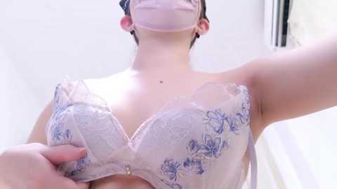 Media: Video of a woman with fair skin wearing a pink surgical mask, a white lace bra with blue floral embroidery, and a black strapless top. She has short, dark hair and is indoors in a bright, white-lit room.