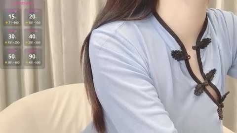 Media: A video of an East Asian woman with long, straight brown hair, wearing a light blue, traditional Chinese blouse with black trim and floral buttons, sitting on a beige chair. Background includes a beige curtain.