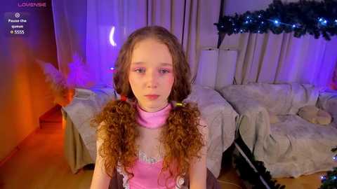 Media: Video of a young girl with curly brown hair styled in pigtails, wearing a pink dress and choker, standing in a cozy, dimly lit living room with purple and orange lighting, a plush teddy bear, and a Christmas tree in the background.