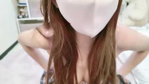 Media: Video of a woman with long, straight brown hair, wearing a white surgical mask, kneeling on a white floor. Background shows a white cabinet with books and a beige couch.